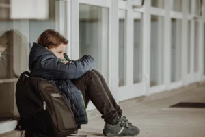 Un niño solitario con mochila sentado en el suelo de un colegio, representando el sentimiento de aislamiento que puede generar el acoso escolar.